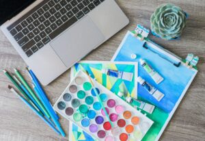 Top-down view of a creative workspace featuring watercolor paints, brushes, and a laptop.