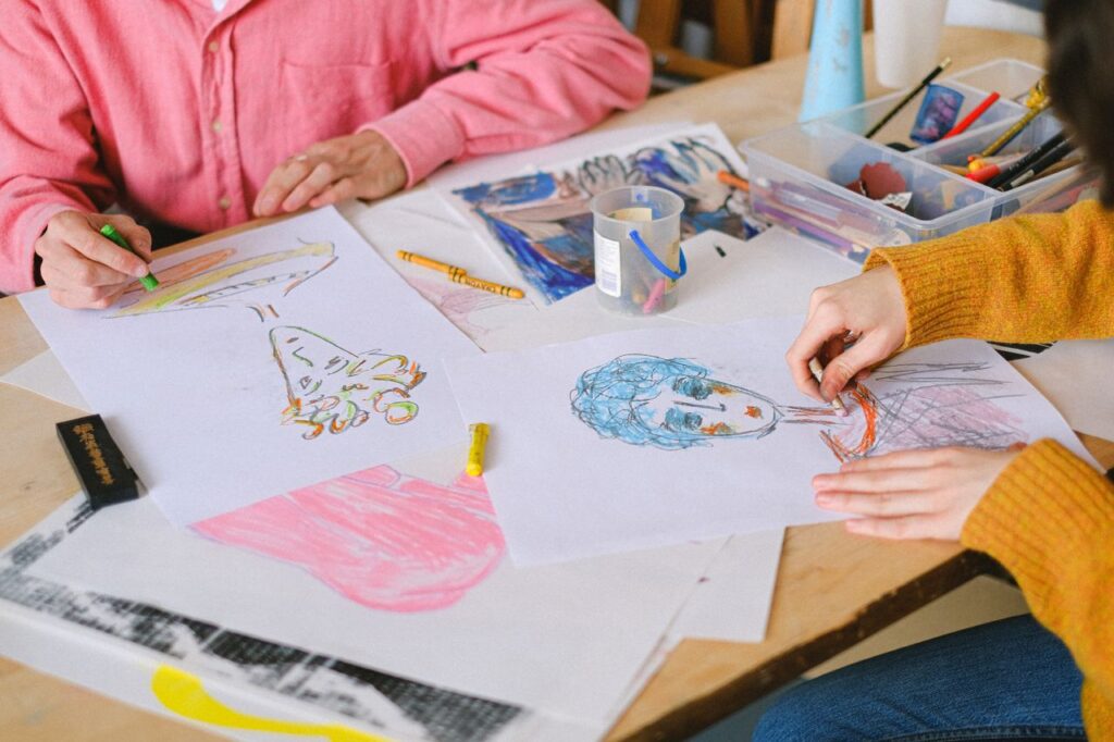 From above of talented artists drawing pictures with colorful crayons while sitting at table and creating paintings in art studio